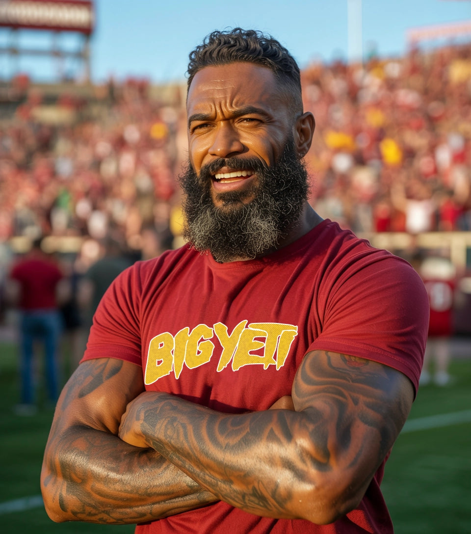 A guy wearing a red t-shirt with the words Big Yeti across the chest. The man is standing cross arm with fans in the background.